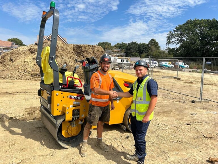 Two construction workers shaking hands on a dirt site next to yellow compacting machinery under blue sky